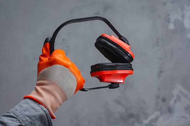 A hand in an orange glove holds red protection headphones gear against a textured grey background. Concept construction safety and protection